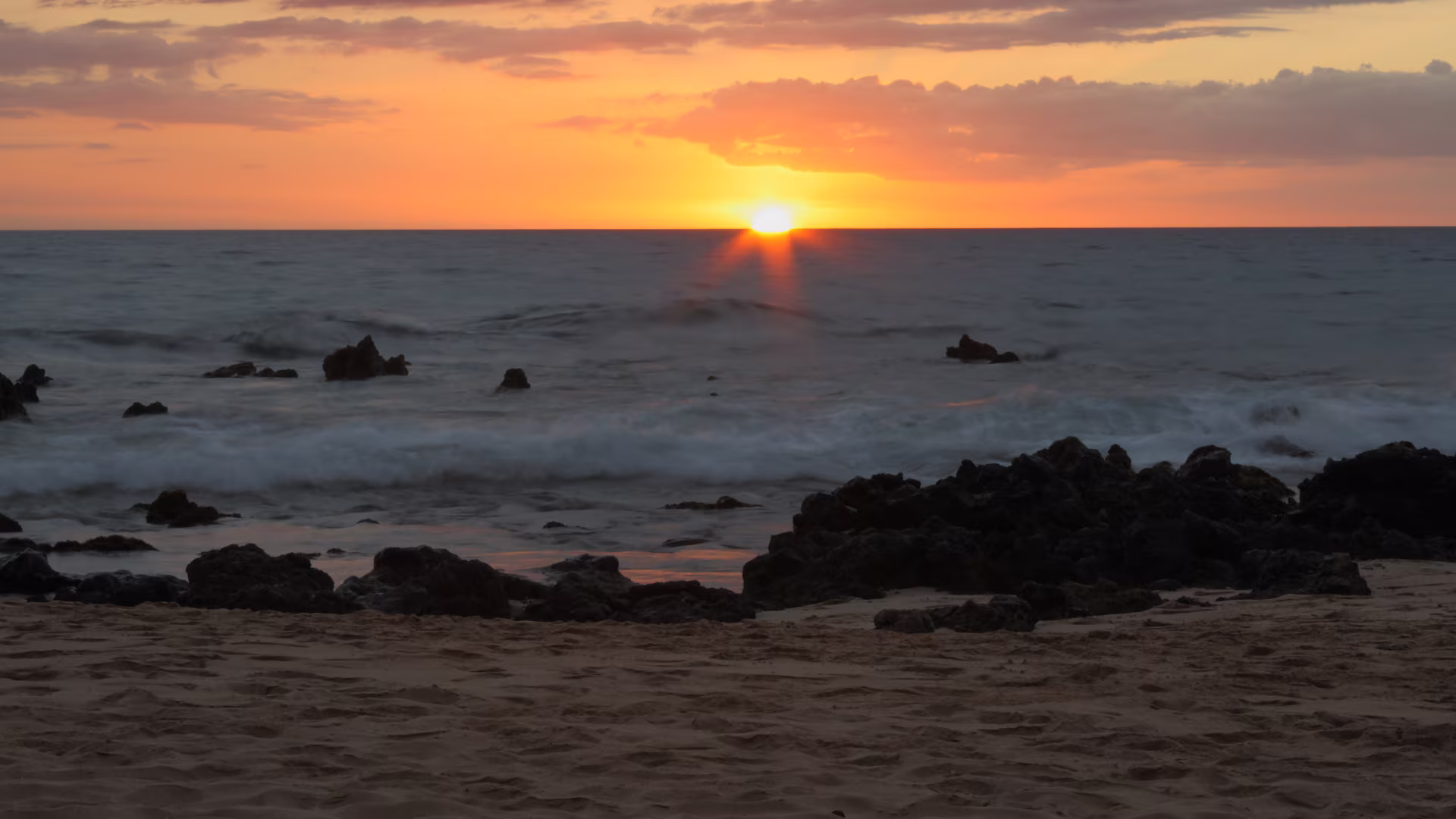 Sunset over the ocean at Keawakapu Beach, Kihei, Maui, with golden sand and lava rock in the foreground