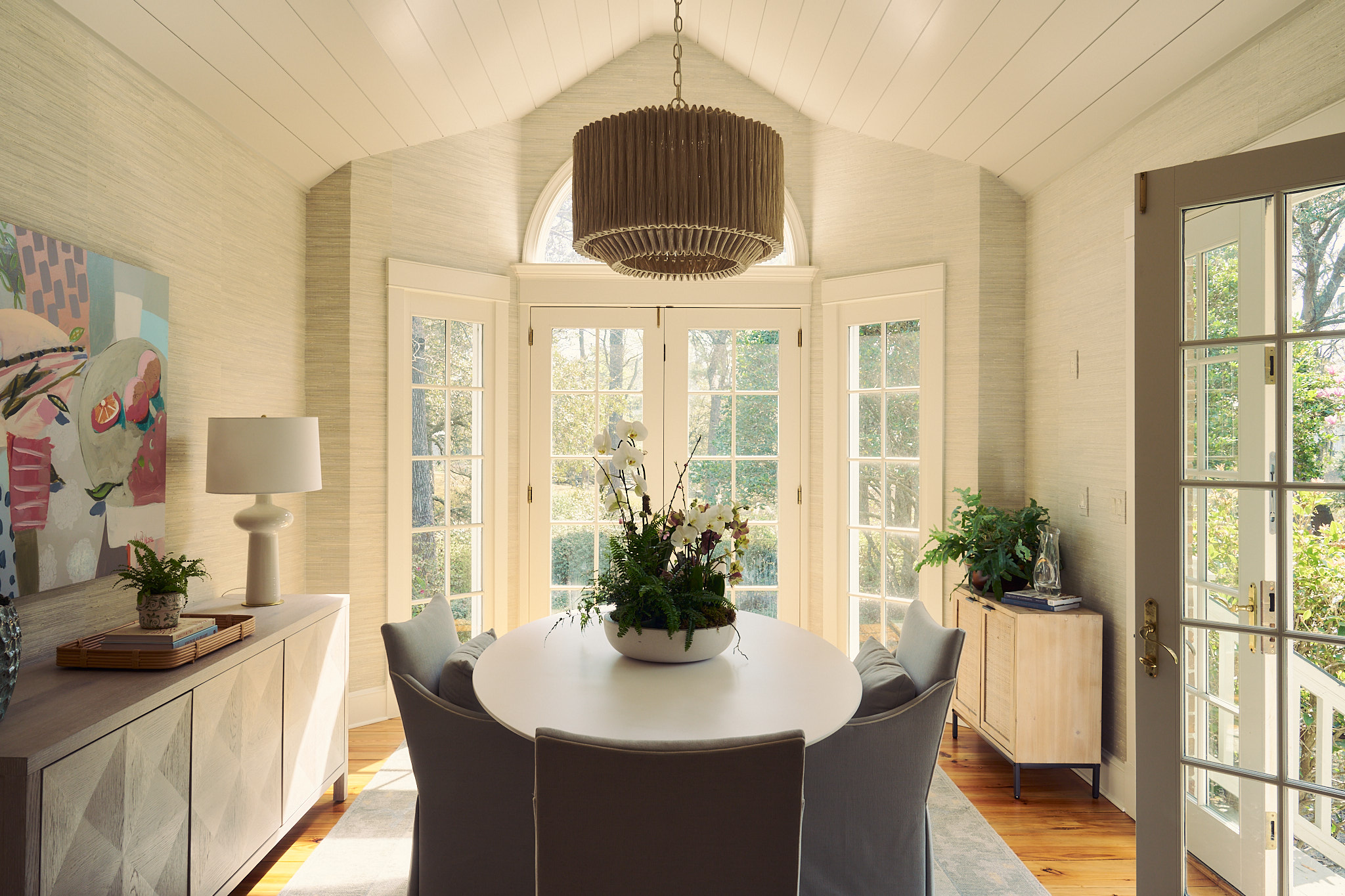 Sunlit breakfast room in a renovated Lowcountry home featuring vaulted shiplap ceiling, round dining table with upholstered chairs, woven statement pendant, soft neutral palette, and French doors opening to garden views in Hobcaw Creek, Charleston South Carolina