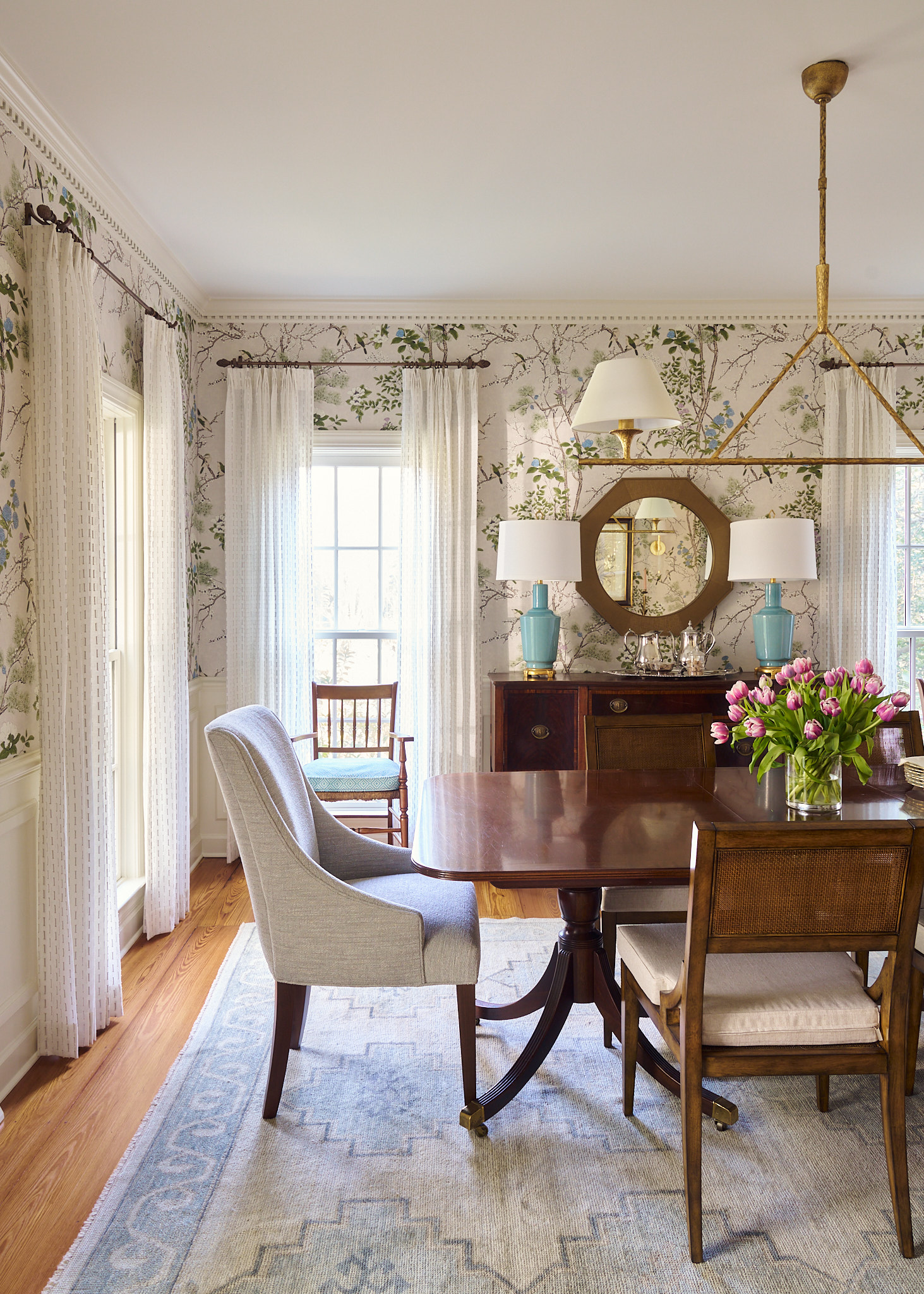 Light-filled Charleston dining room with botanical wallpaper, wood dining table, upholstered and cane-back chairs, brass chandelier, and styled sideboard with blue ceramic lamps and fresh tulips for a layered, timeless interior.