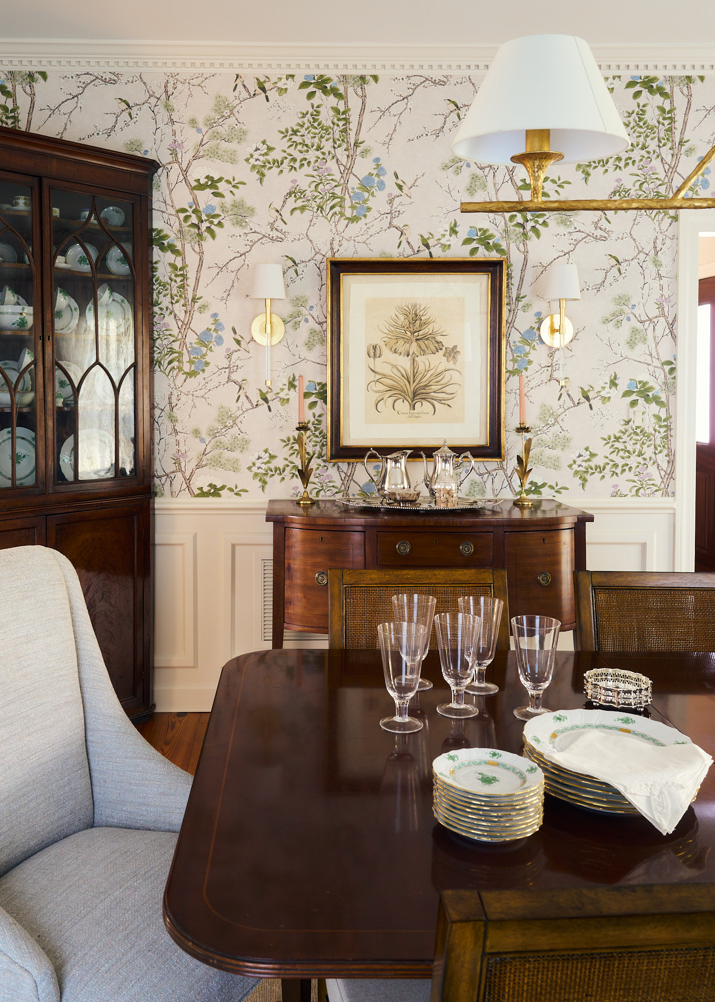 Traditional Charleston dining room with botanical wallpaper, dark wood dining table and sideboard, antique china cabinet, brass sconces, and styled tabletop with glassware and china for a layered, timeless interior.