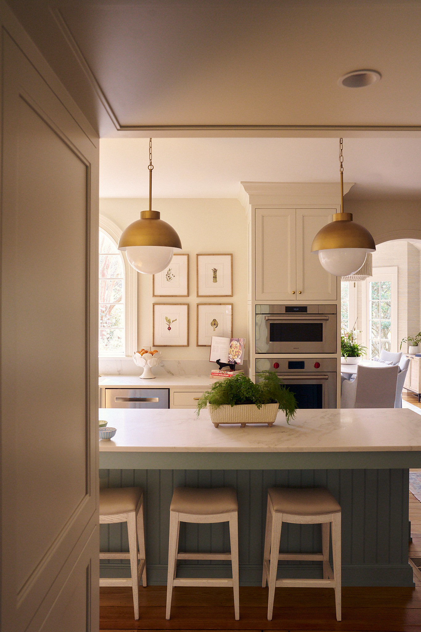 Charleston kitchen with marble island and soft blue paneled base, paired with brass dome pendant lights, inset cabinetry, integrated appliances, and open sightlines to a light-filled dining room beyond.
