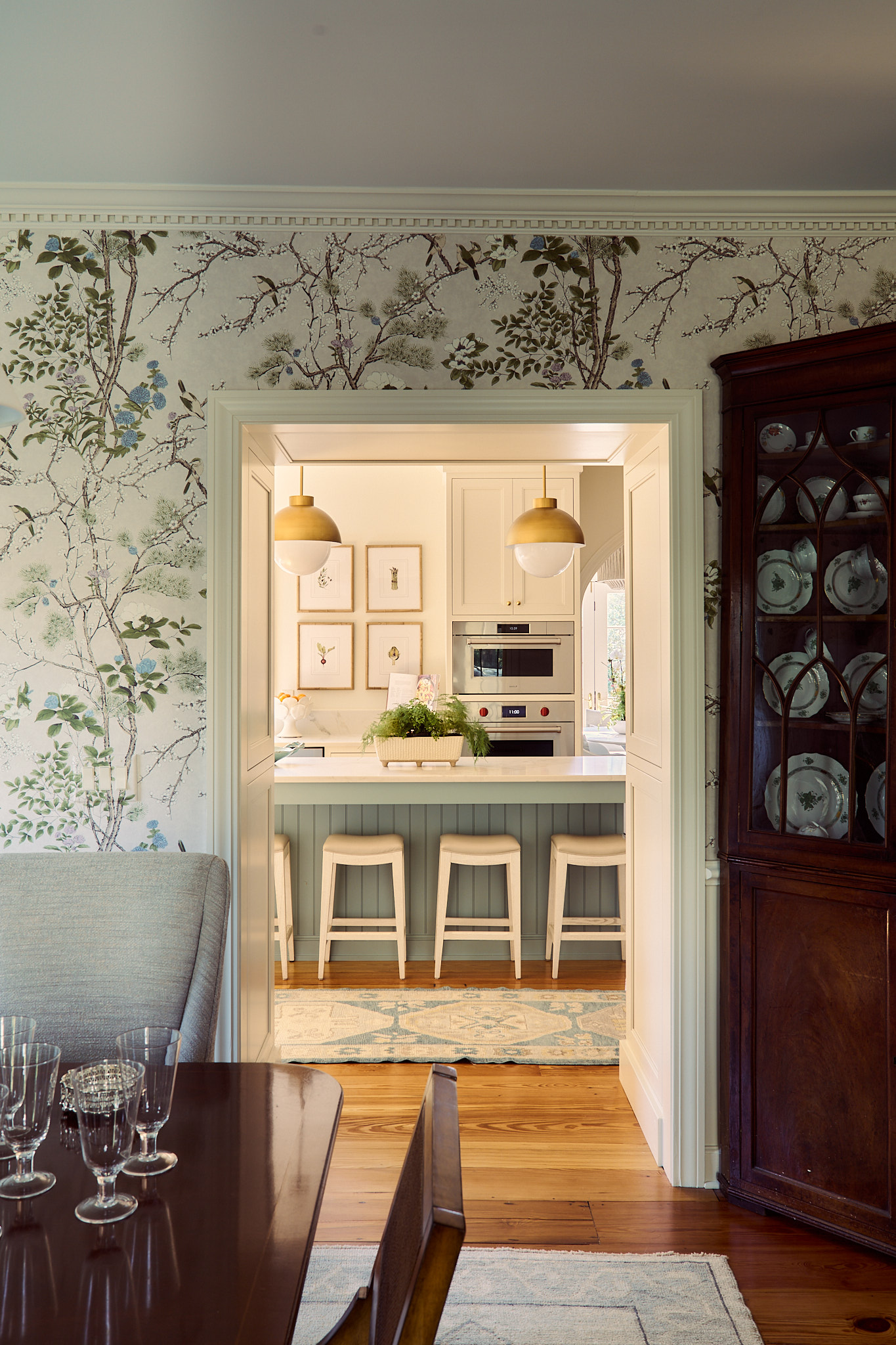Dining room with botanical wallpaper and traditional wood china cabinet, framing a view into a Charleston kitchen with a soft blue island, marble countertop, brass pendant lighting, and warm, light-filled interiors.