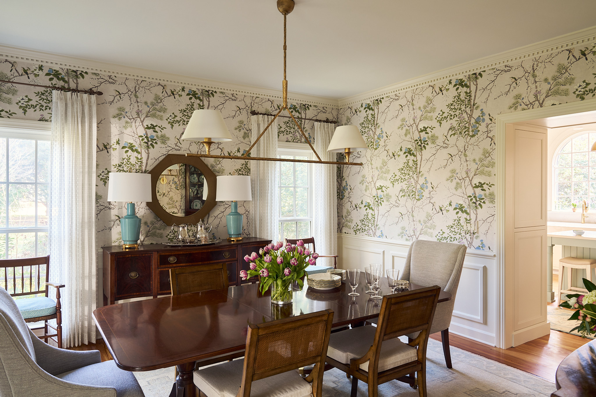 Traditional dining room in a renovated Hobcaw Creek home featuring botanical wallpaper, antique wood dining table, brass chandelier, blue ceramic lamps, and soft linen drapery creating a bright, layered Lowcountry interior in Charleston South Carolina