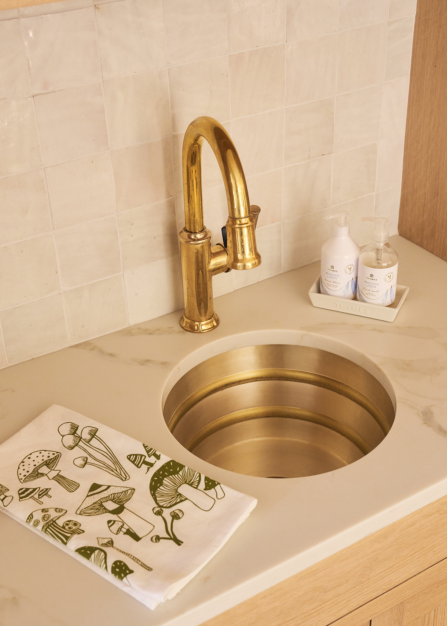 Warm brass sink and faucet set into a marble countertop with zellige tile backsplash, styled with soap dispensers and a mushroom print linen, showcasing layered texture and refined details in a Charleston kitchen.