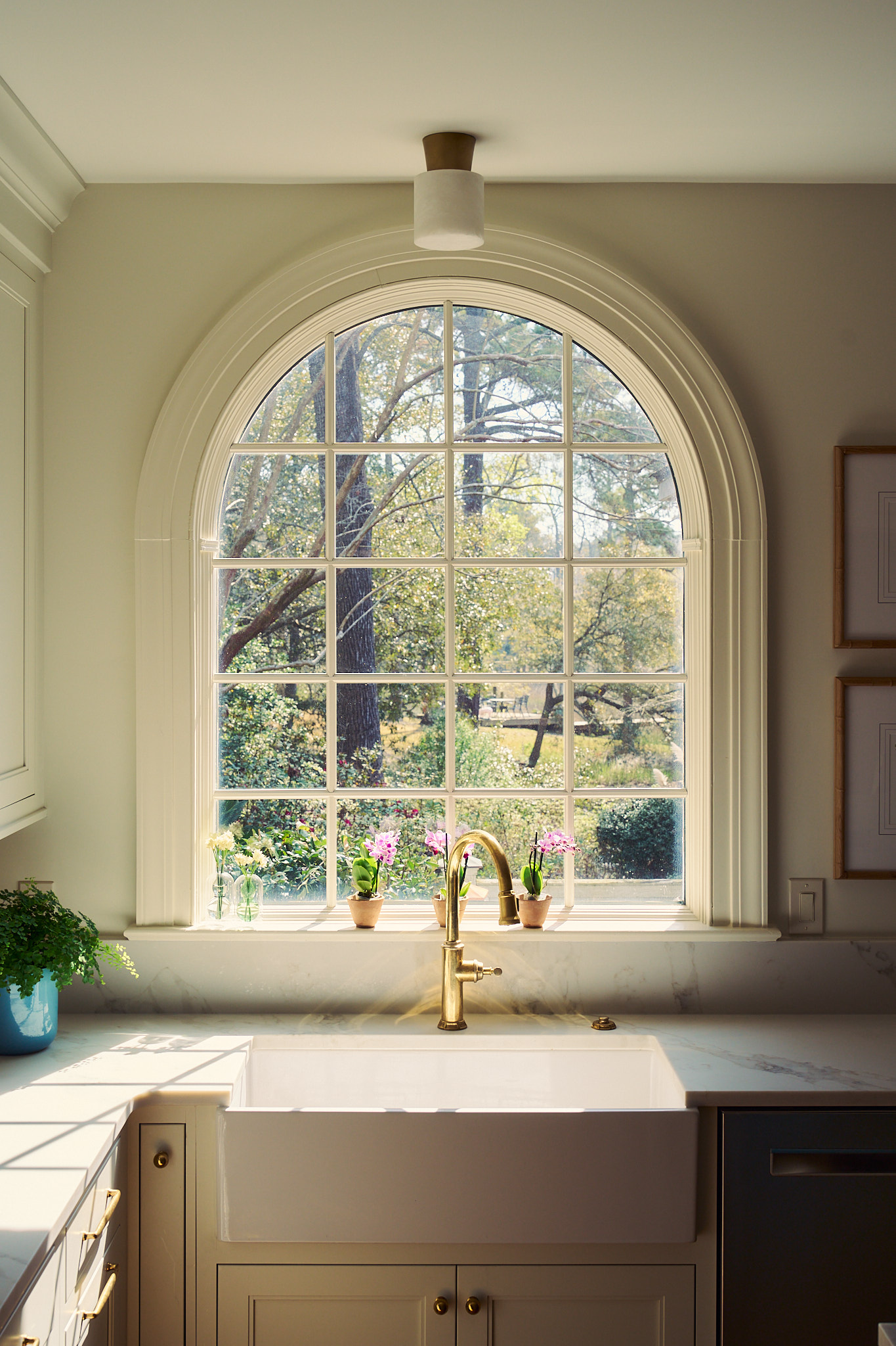 Arched kitchen window above a farmhouse sink with brass faucet, marble countertops, and soft neutral cabinetry, styled with small potted plants and overlooking a lush Charleston garden filled with natural light.