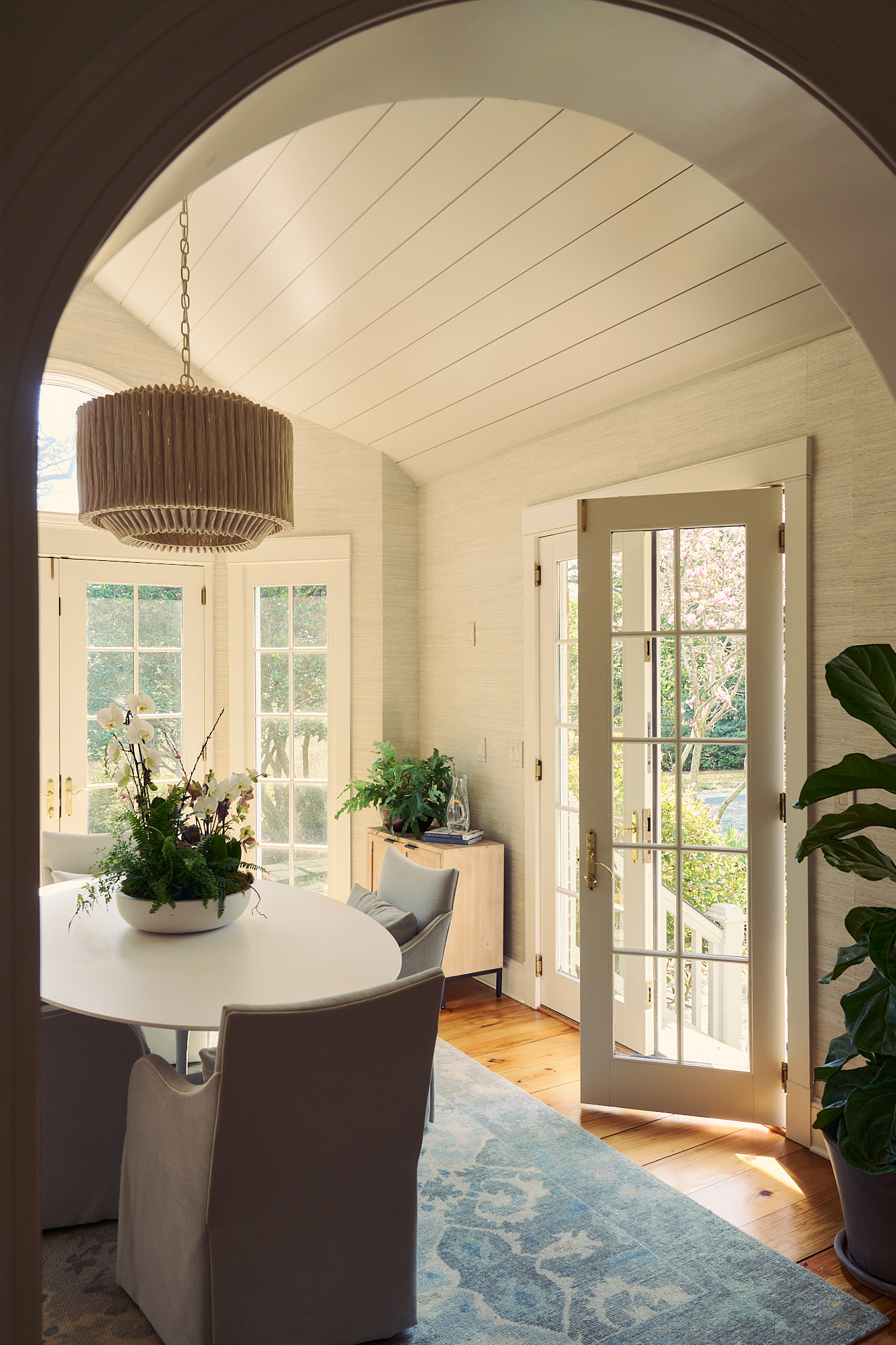 Arched entry view into a bright Charleston dining room with round table, upholstered chairs, textured grasscloth walls, statement pleated pendant, and French doors opening to a sunlit garden.Arched kitchen window above a farmhouse sink with brass faucet, m