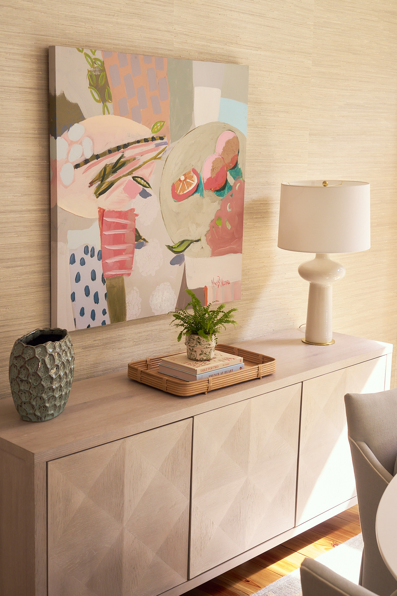 Light wood sideboard with geometric panel detailing styled beneath a colorful abstract painting, featuring a ceramic table lamp, woven tray with books and greenery, and textured grasscloth wallpaper in a warm, layered Charleston interior.