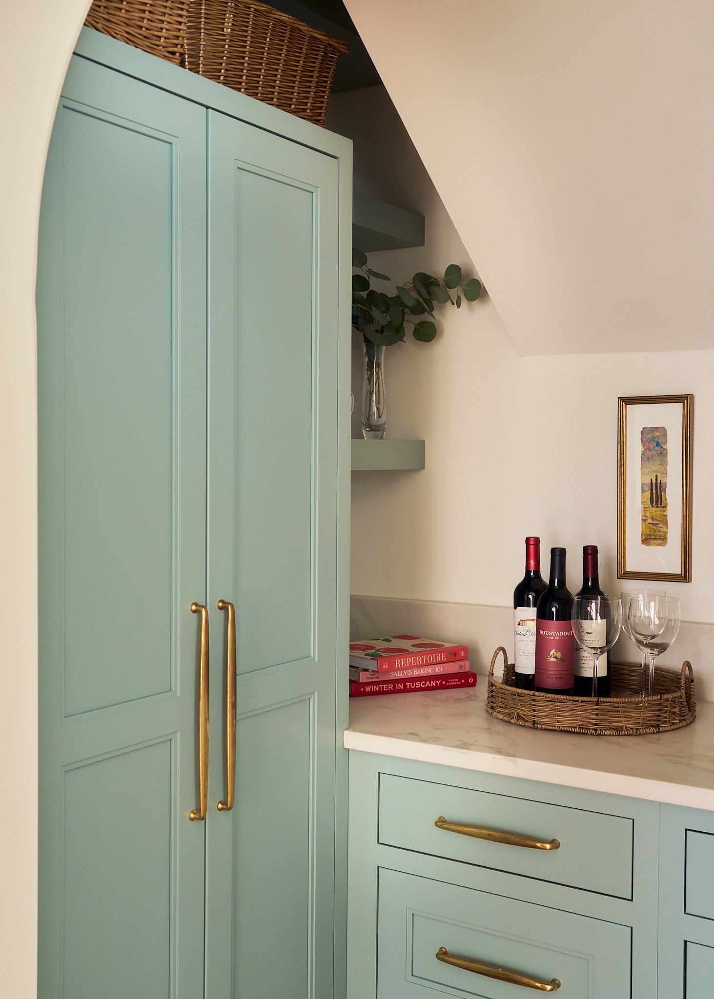 Soft blue custom cabinetry with brass hardware in a Charleston kitchen bar area, styled with marble countertop, woven tray with wine bottles and glasses, open shelving with greenery, and sloped ceiling detail for a cozy built-in nook.
