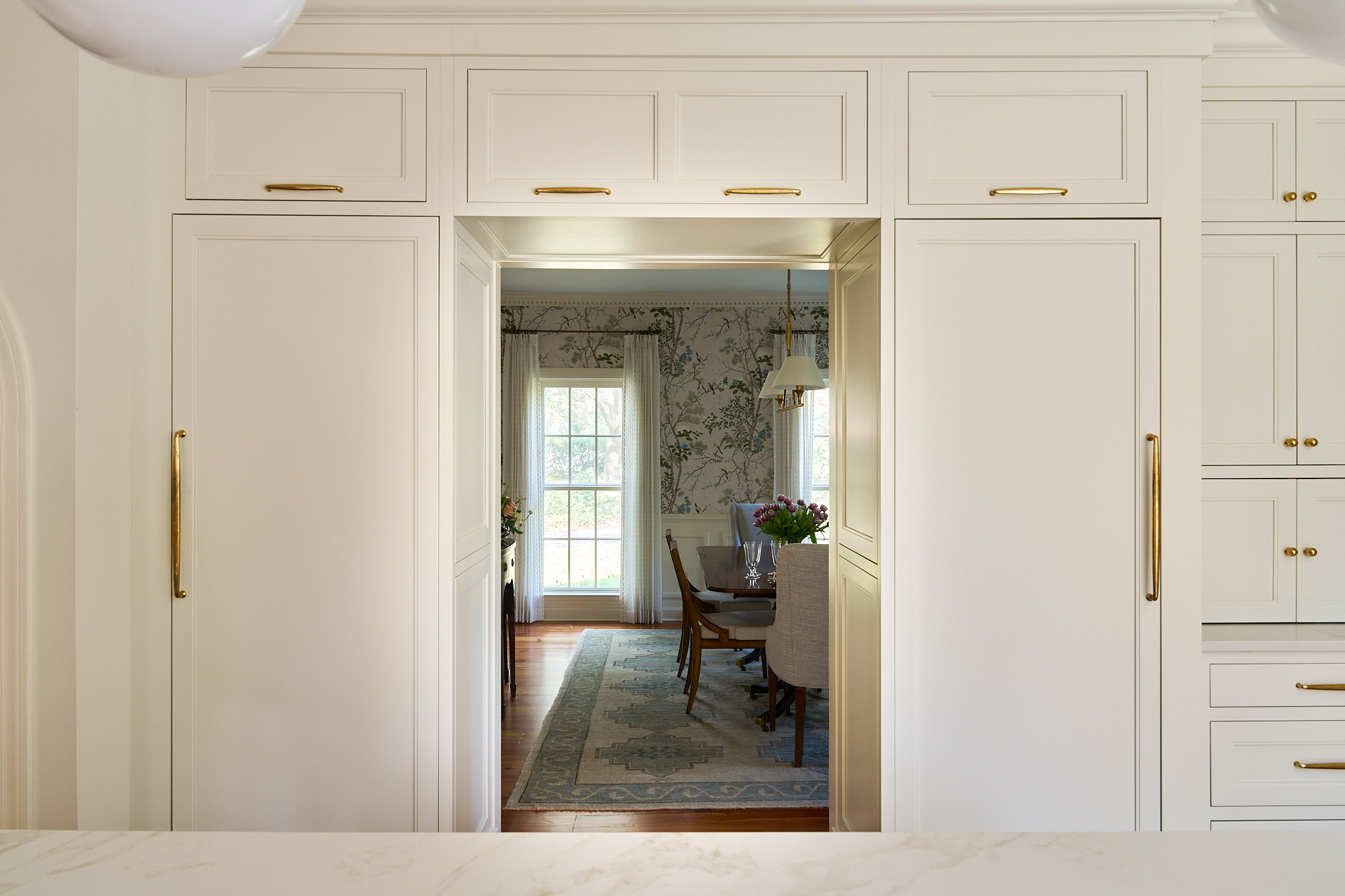 Custom kitchen cabinetry with integrated paneled refrigerator and brass hardware framing a view into a traditional dining room with botanical wallpaper, creating a seamless, light filled transition in a Hobcaw Creek home in Charleston South Carolina