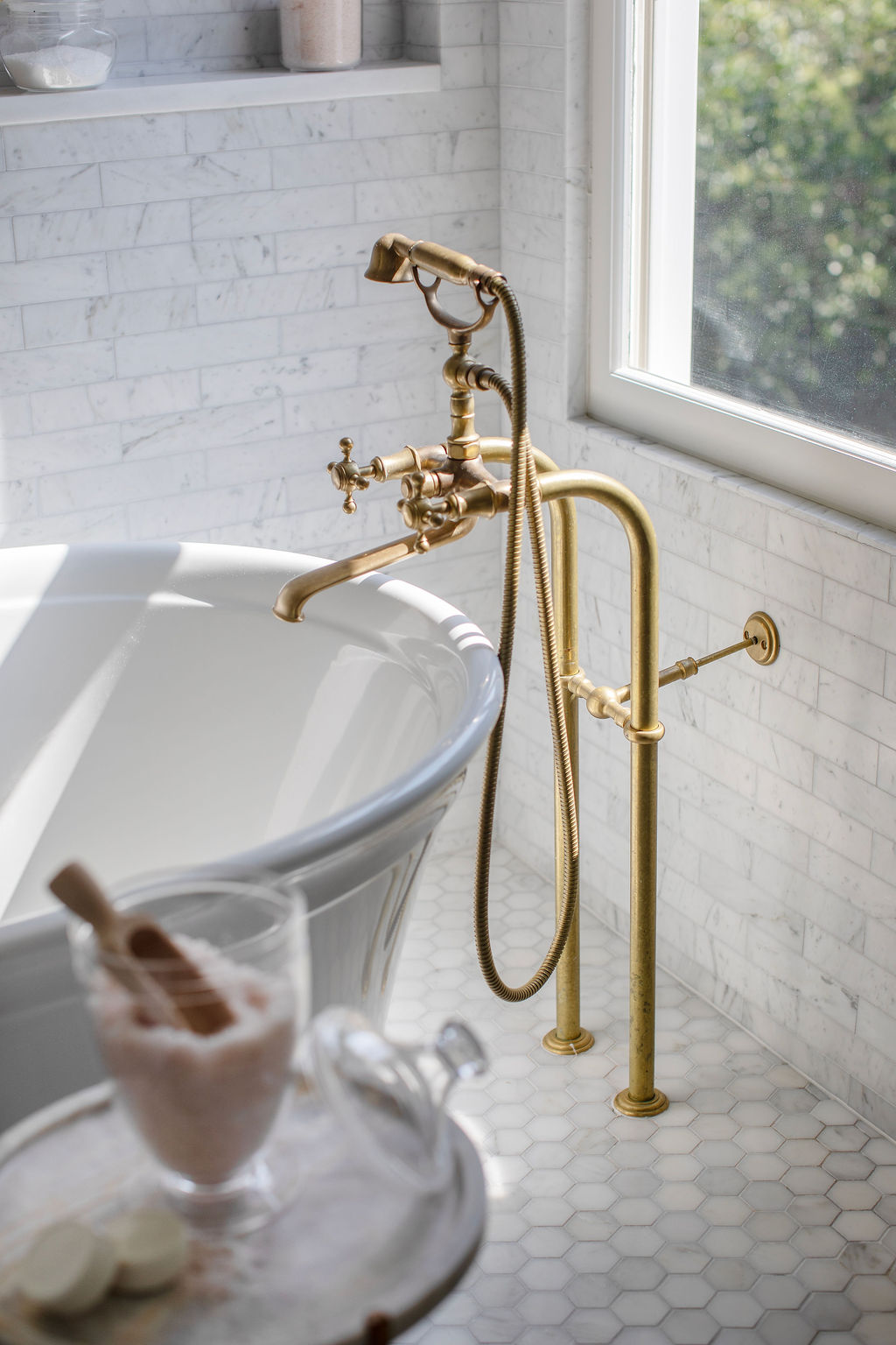 Freestanding soaking tub with unlacquered brass floor-mounted tub filler, marble tile walls, and abundant natural light. Designed by PWD Studio, this spa-inspired bathroom combines timeless luxury and classic craftsmanship.