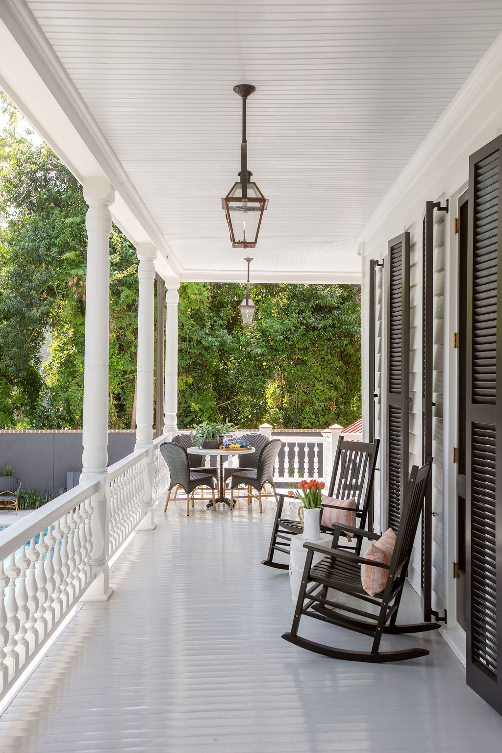 Classic Charleston piazza featuring white columns, black shutters, beadboard ceilings, lantern pendants, and black rocking chairs overlooking a lush garden. Designed by PWD Studio, this timeless Southern porch blends historic architecture, outdoor living, 