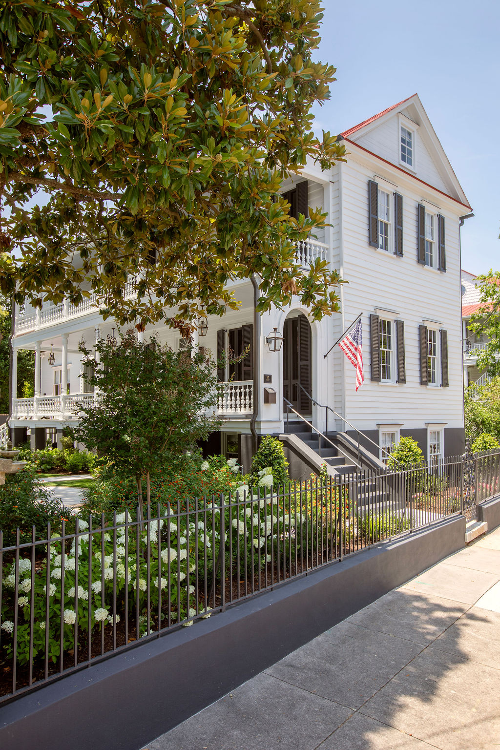 Elegant Charleston single house with expansive piazzas, traditional shutters, lush landscaping, and wrought iron fencing. Designed by PWD Studio, the property combines historic preservation with sophisticated outdoor living.