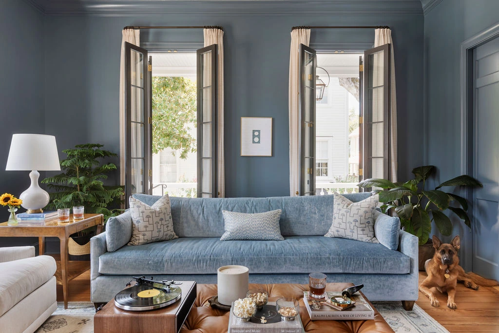 Elegant blue living room interior with moody painted walls, a light blue velvet sofa, French casement windows, warm wood accents, layered neutral textiles, and 