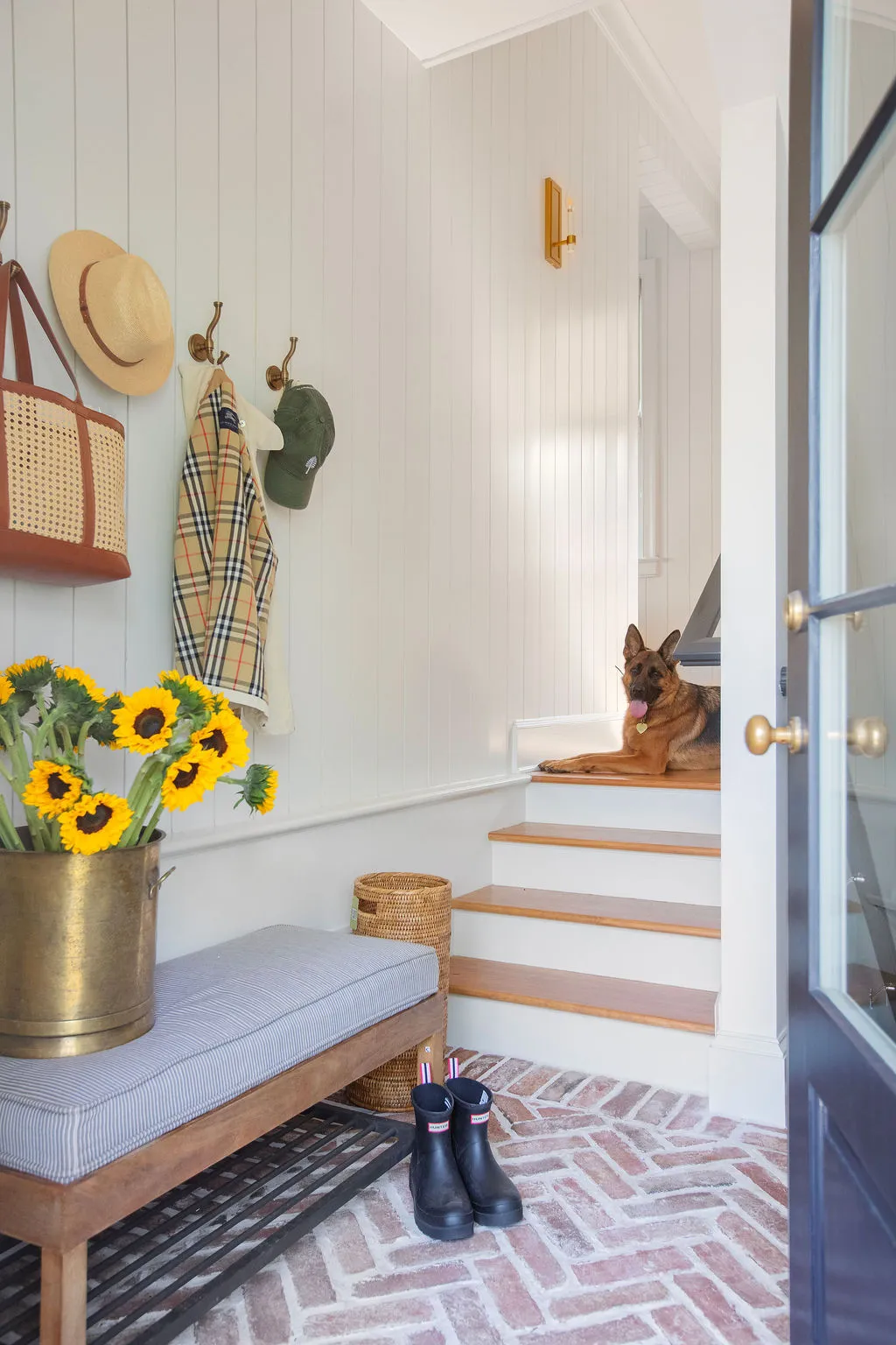 Inviting mudroom entry with reclaimed brick flooring, shiplap walls, custom bench seating, brass hooks, and a German Shepherd resting on the staircase. Designed by PWD Studio, the space blends practical family living with timeless Lowcountry charm.