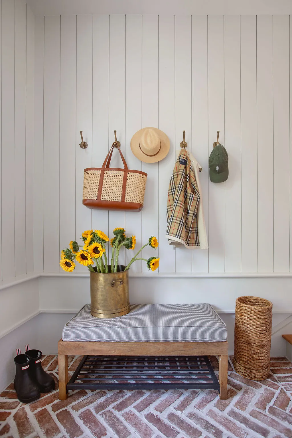 Charming mudroom featuring reclaimed brick floors, custom bench seating, woven accessories, brass hooks, and fresh sunflowers. Designed by PWD Studio, the functional entry combines classic Charleston architecture with tailored storage and everyday elegance