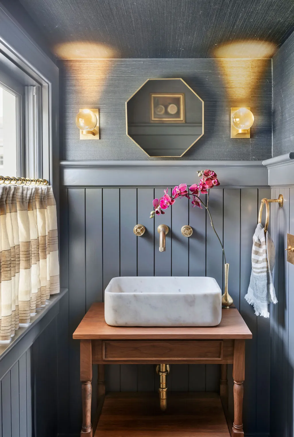 Sophisticated powder room featuring dark grasscloth wallpaper, painted millwork, a marble vessel sink, warm walnut vanity, and unlacquered brass fixtures. Designed by PWD Studio, the space combines dramatic color, rich texture, and timeless luxury.