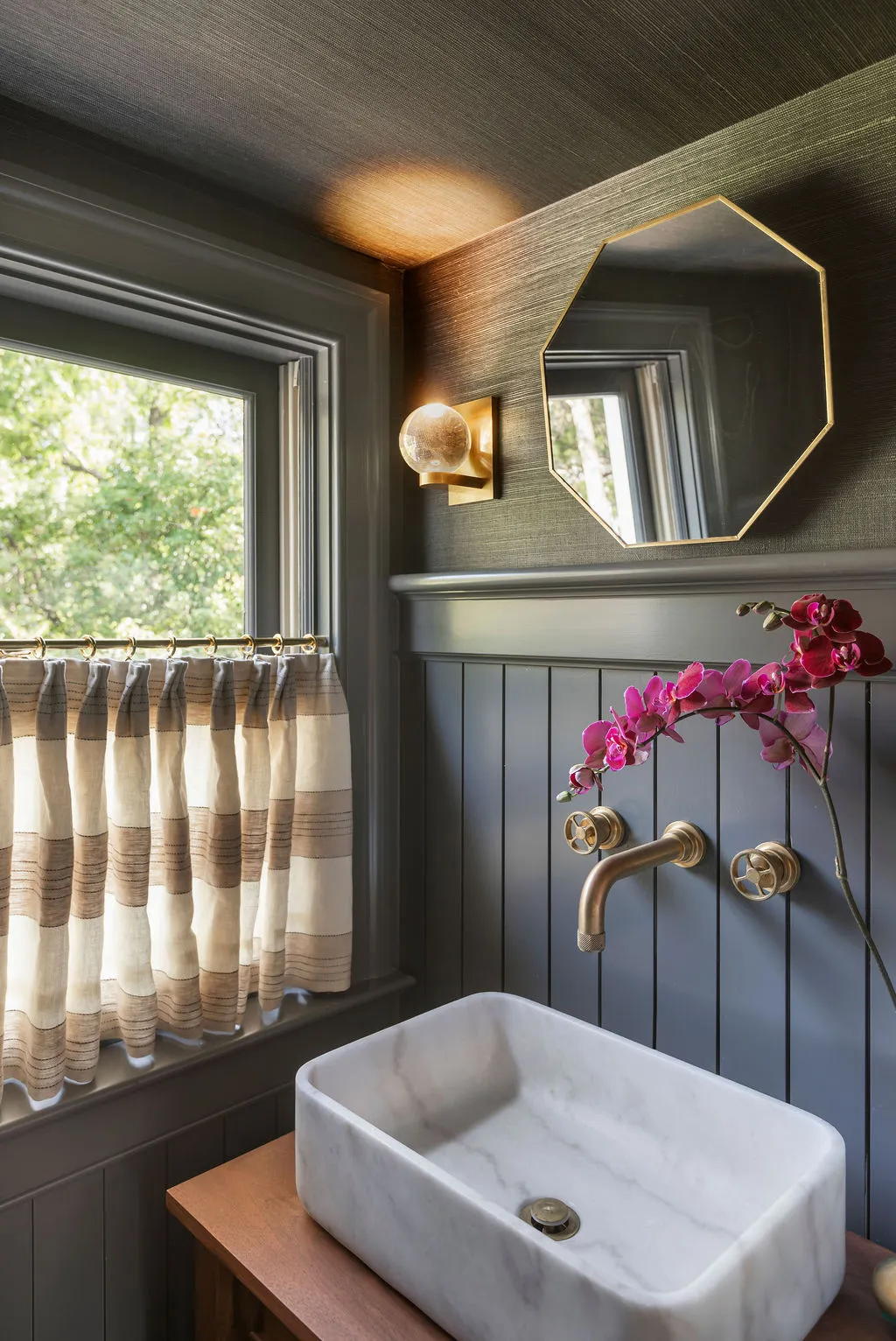 Elegant powder room vanity with a marble vessel sink, brass wall-mounted faucet, octagonal mirror, and textured grasscloth walls. Designed by PWD Studio, this intimate space blends moody sophistication, custom detailing, and refined Charleston style.