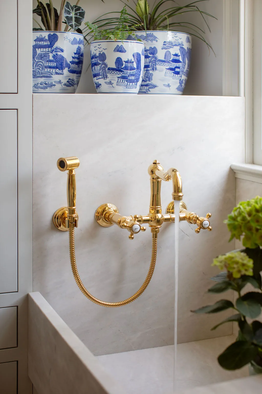 Luxury marble farmhouse sink with unlacquered brass wall-mounted faucet and blue-and-white porcelain planters. Designed by PWD Studio, this custom pantry detail highlights artisan materials, classic fixtures, and elegant Charleston interior design.