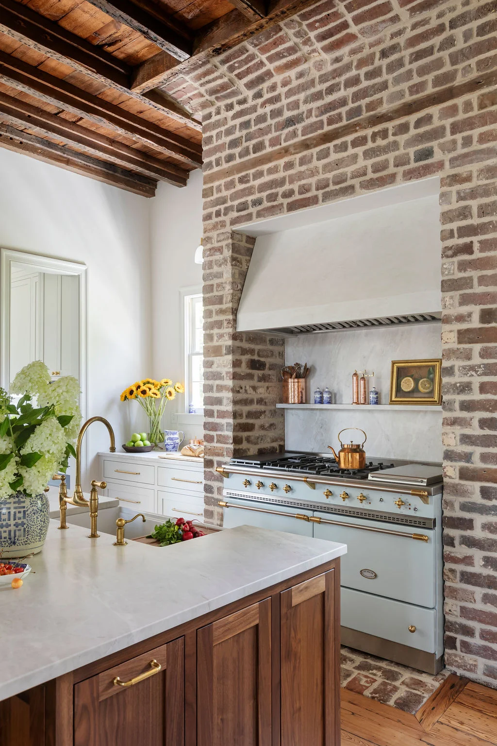 Luxury kitchen featuring exposed brick walls, reclaimed wood ceilings, a custom walnut island, marble countertops, and a professional range framed by a plaster hood. Designed by PWD Studio, the space blends historic Charleston character with timeless craft