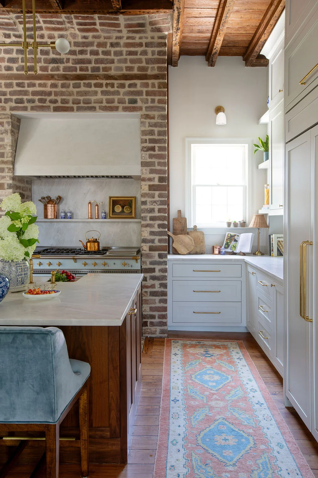 Luxury kitchen featuring exposed brick walls, reclaimed wood ceilings, a custom walnut island, marble countertops, and a professional range framed by a plaster hood. Designed by PWD Studio, the space blends historic Charleston character with timeless craft
