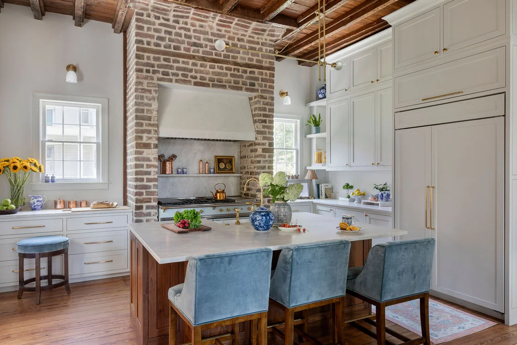 Luxury historic kitchen with exposed brick walls, reclaimed wood beam ceilings, custom white cabinetry, marble island, brass fixtures, and contemporary traditional design blending Charleston charm with modern functionality.
