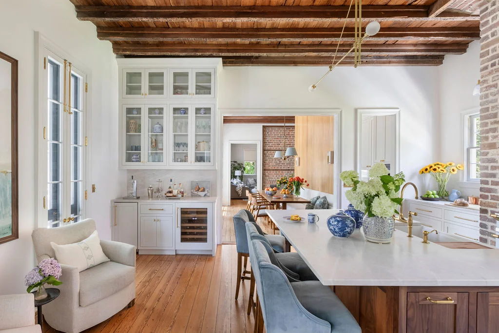 Historic Charleston kitchen with exposed wood beam ceilings, original brick walls, marble waterfall island, custom cabinetry, brass fixtures, and contemporary traditional interiors blending preserved architecture with refined modern living.