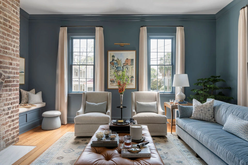 Moody blue drawing room with color drenched walls, velvet sofa, French casement windows, warm wood accents, layered textiles, and contemporary traditional Charleston interior design.