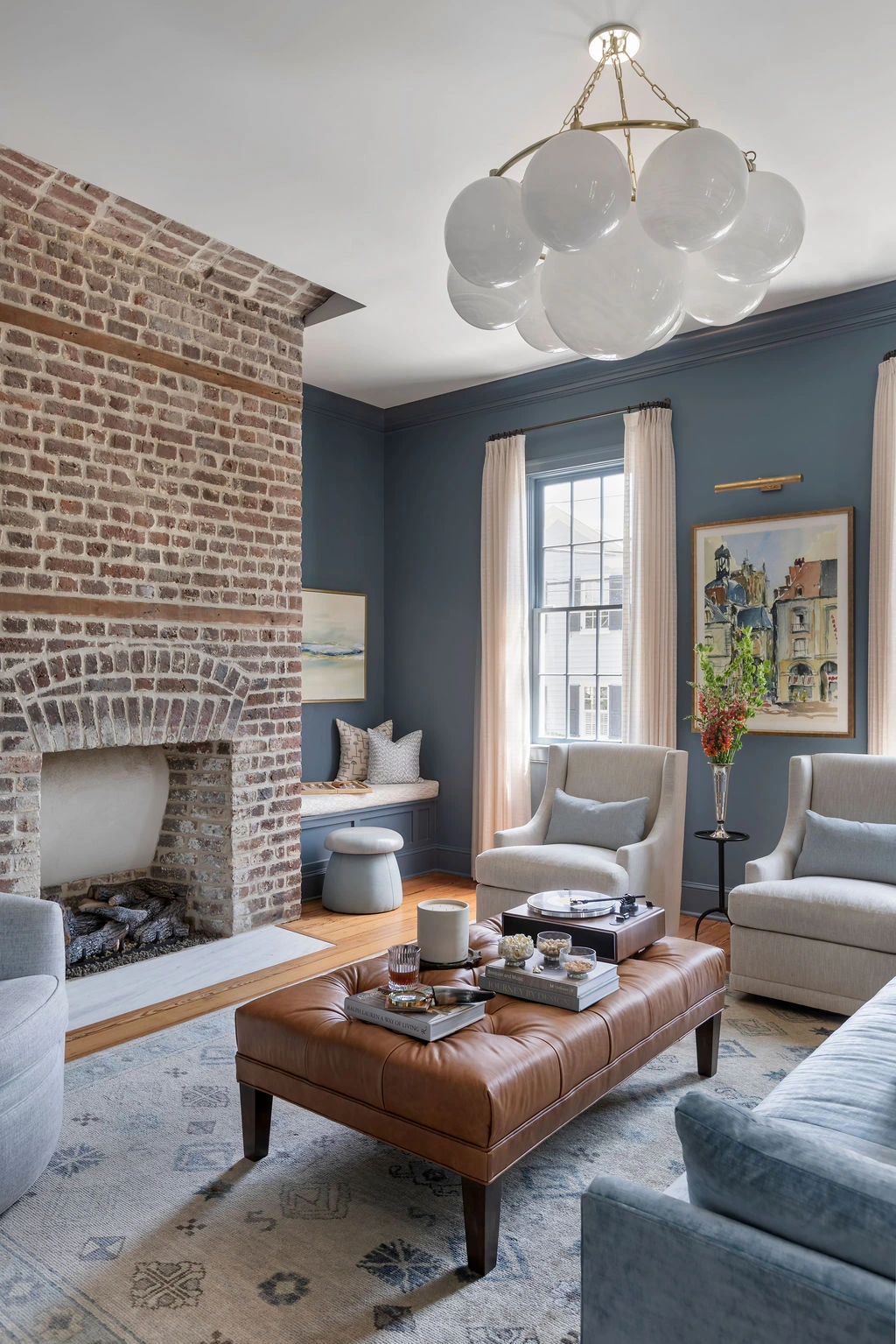 Elegant Charleston living room with exposed brick fireplace, deep blue walls, custom upholstery, leather ottoman, and statement globe chandelier. Designed by PWD Studio, the space combines historic charm, layered textures, and sophisticated Lowcountry inte