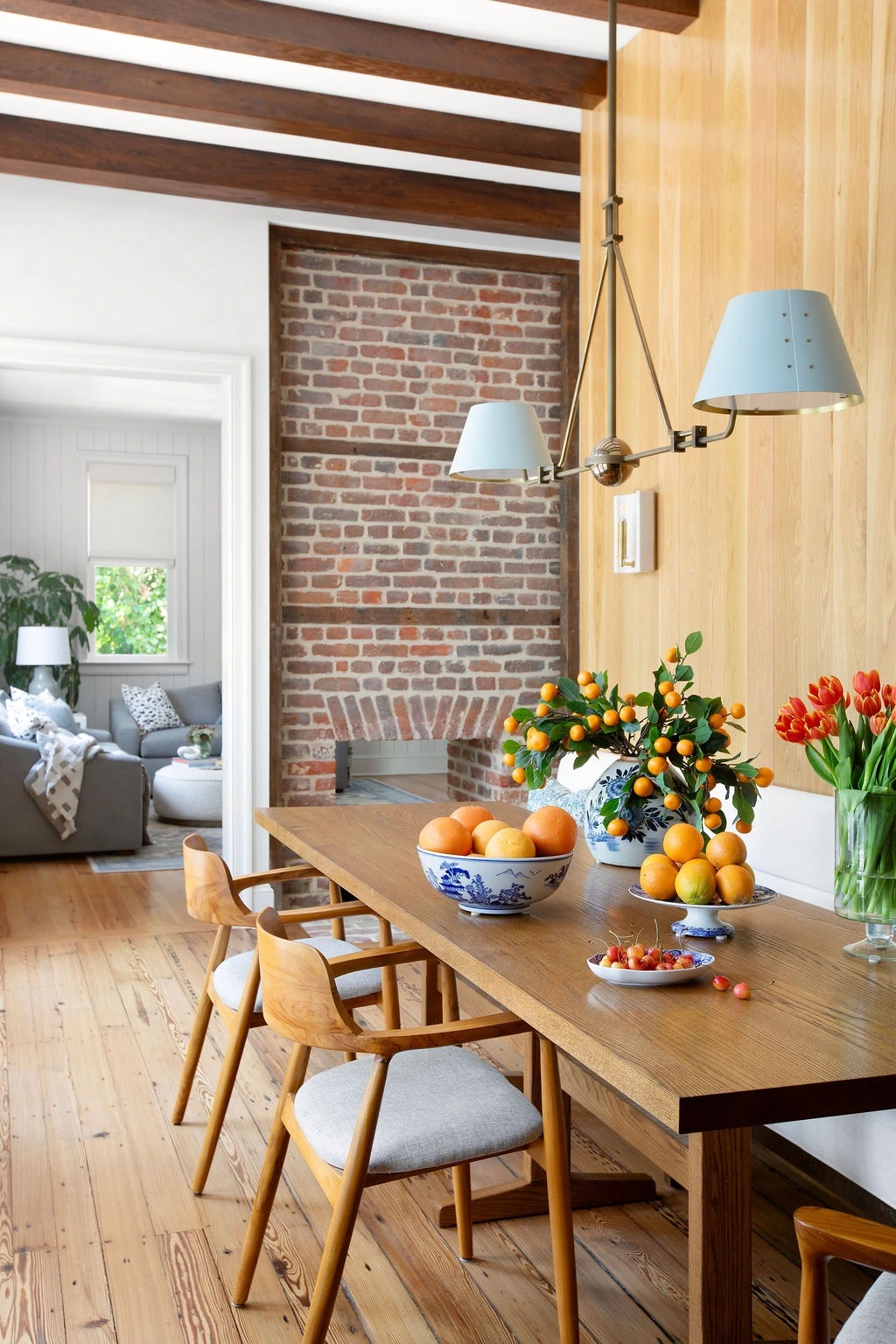 Sophisticated dining room featuring a custom white oak dining table, sculptural wood dining chairs, exposed brick fireplace, and warm wood ceiling beams. Designed by PWD Studio, the space blends historic Charleston character with refined craftsmanship, nat