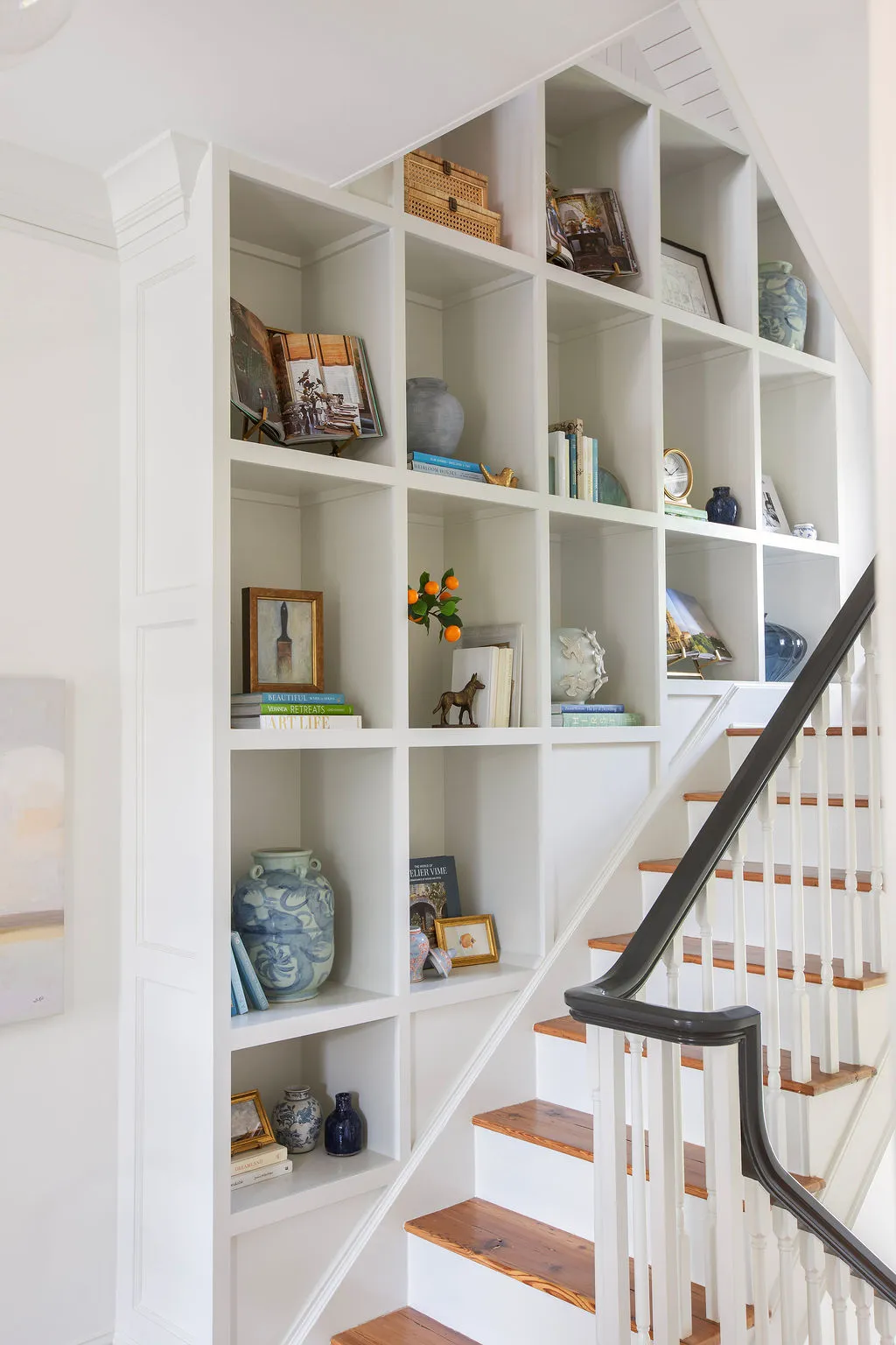 Custom floor-to-ceiling staircase bookcase featuring curated artwork, collected antiques, blue-and-white ceramics, and decorative styling. Designed by PWD Studio, this Charleston home showcases bespoke millwork, functional storage, and timeless interior ar