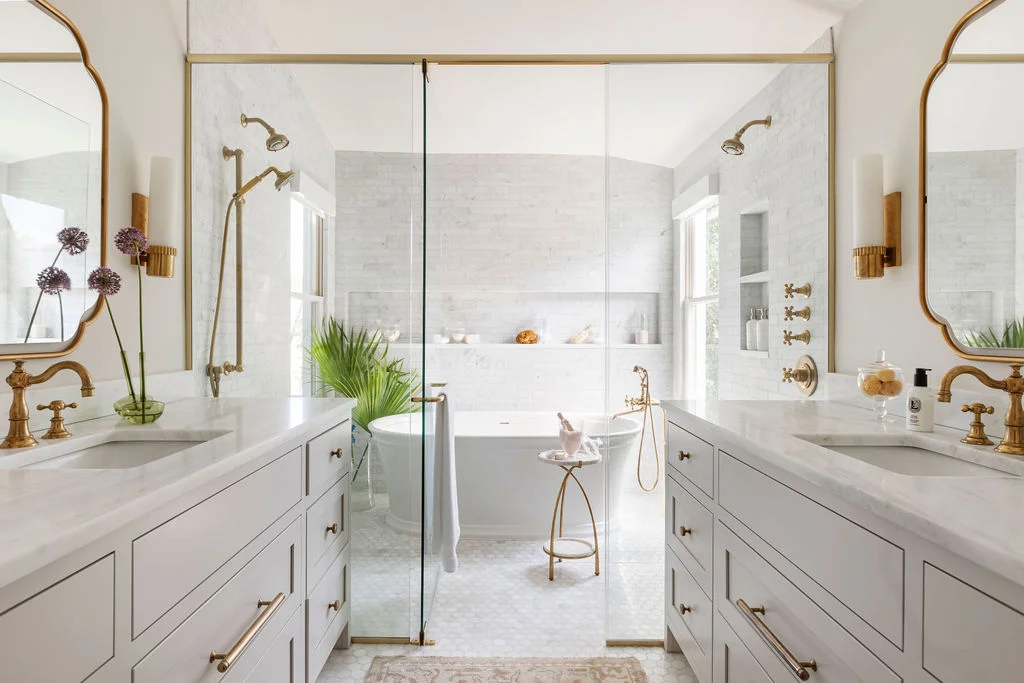 Luxury white marble bathroom with dual custom vanities, unlacquered brass fixtures, glass shower enclosure, freestanding soaking tub, and timeless contemporary traditional design with soft natural light.