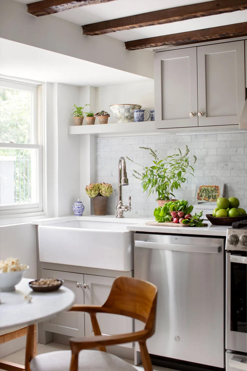 Charming Charleston kitchen featuring a farmhouse sink, marble backsplash, custom gray cabinetry, exposed wood beams, and open shelving styled with greenery and collected ceramics. Designed by PWD Studio, the space blends historic architecture with timeles