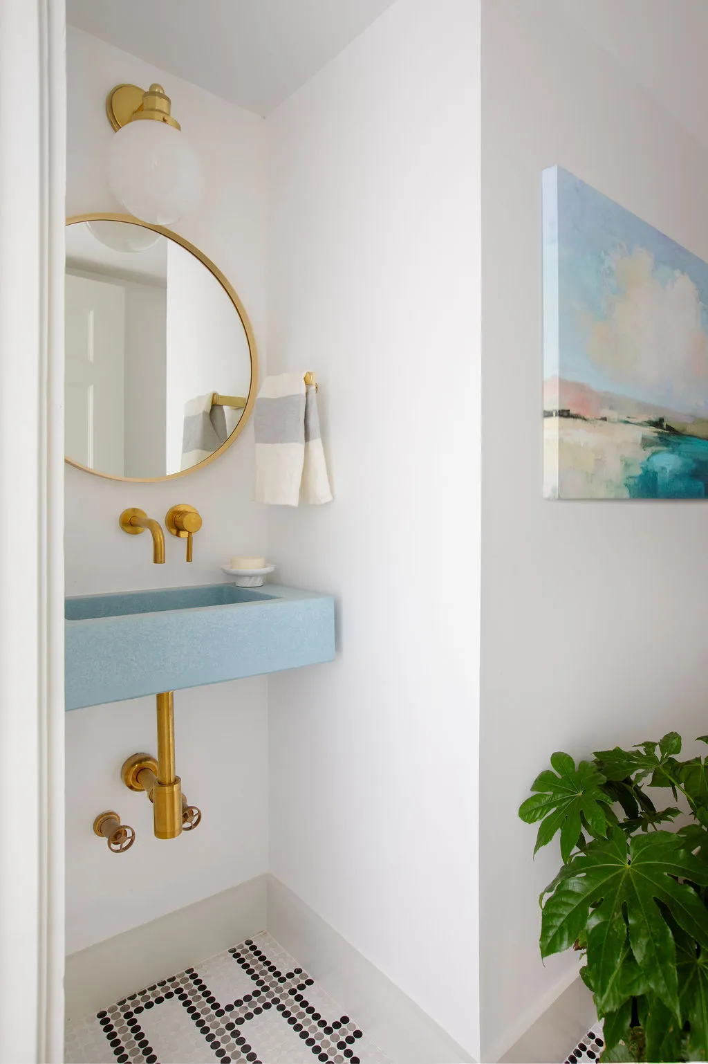 Modern powder room featuring a custom pale blue floating sink, unlacquered brass fixtures, round brass mirror, graphic mosaic tile flooring, and contemporary artwork. Designed by PWD Studio, the space balances playful color, clean lines, and pool bath