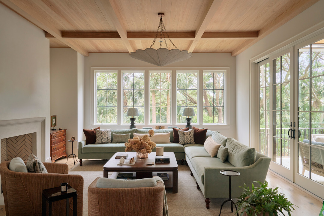 Elegant Kiawah Island living room designed by PWD Studio featuring a custom pale green sectional sofa, warm wood coffered ceiling, white oak flooring, tailored upholstery, and expansive windows overlooking lush coastal landscape. The refined interior blends cottage-inspired comfort with sophisticated Lowcountry design, creating a bright and inviting family gathering space filled with natural light.