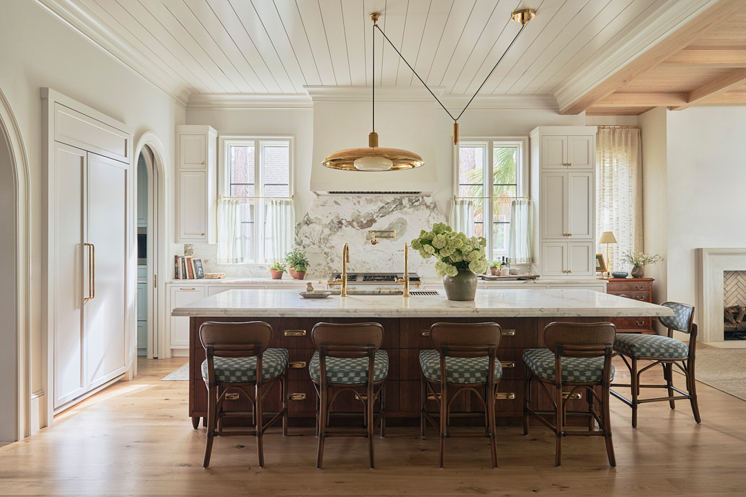 Custom Kiawah Island kitchen featuring a walnut furniture-style island, oversized marble countertops, brass accents, bespoke cabinetry, and a striking natural stone backsplash in a refined coastal home. Luxury Kiawah Island kitchen with a walnut island, marble waterfall countertop, brass plumbing fixtures, custom cabinetry, and a dramatic veined marble backsplash designed for effortless entertaining.