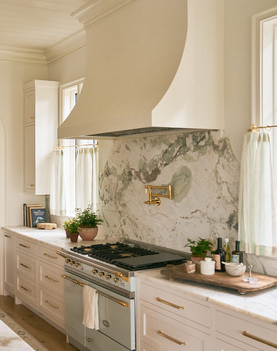 Custom kitchen range wall featuring a sculptural plaster hood, brass pot filler, marble backsplash, and tailored cabinetry blending timeless craftsmanship with modern functionality.