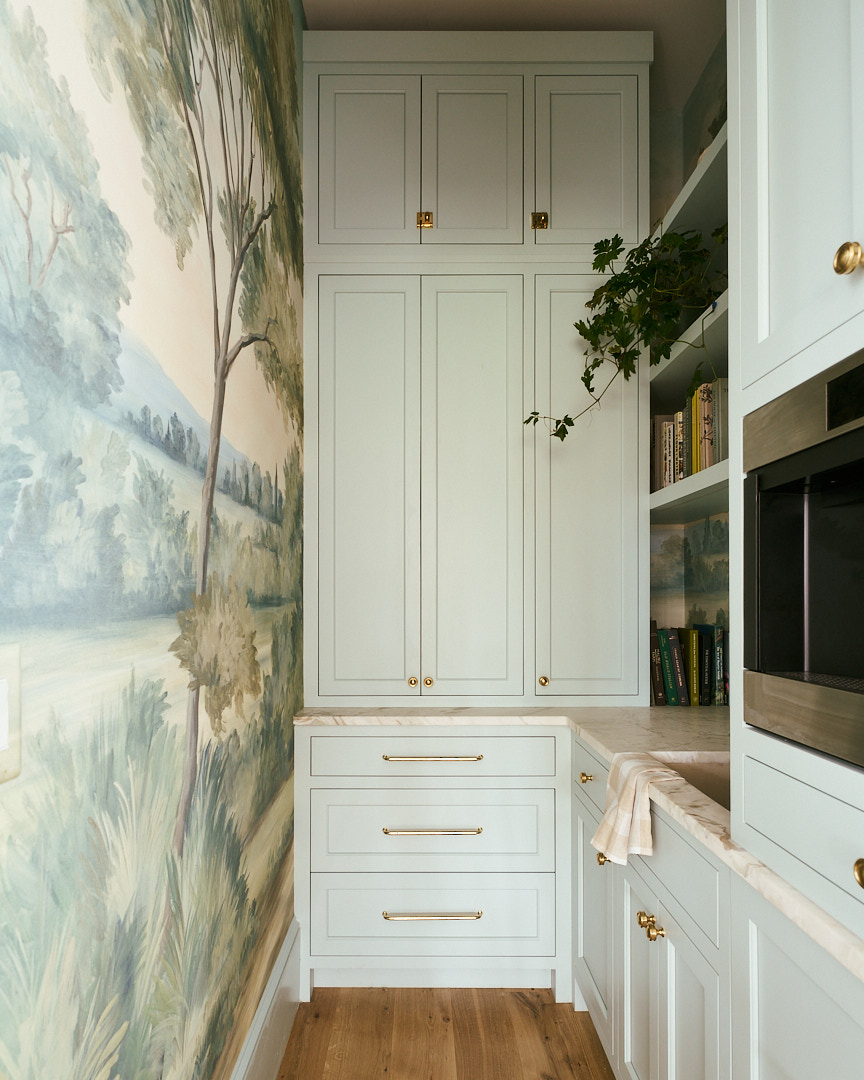 Elegant butler’s pantry featuring pale blue custom cabinetry, brass hardware, marble worktops, integrated storage, and a scenic mural wallcovering inspired by the Lowcountry landscape.
