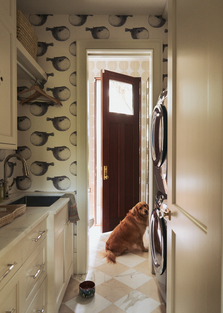 Playful laundry and mudroom with whimsical bird wallpaper, stacked washer and dryer, farmhouse sink, marble flooring, and a golden retriever in a custom Kiawah Island home.