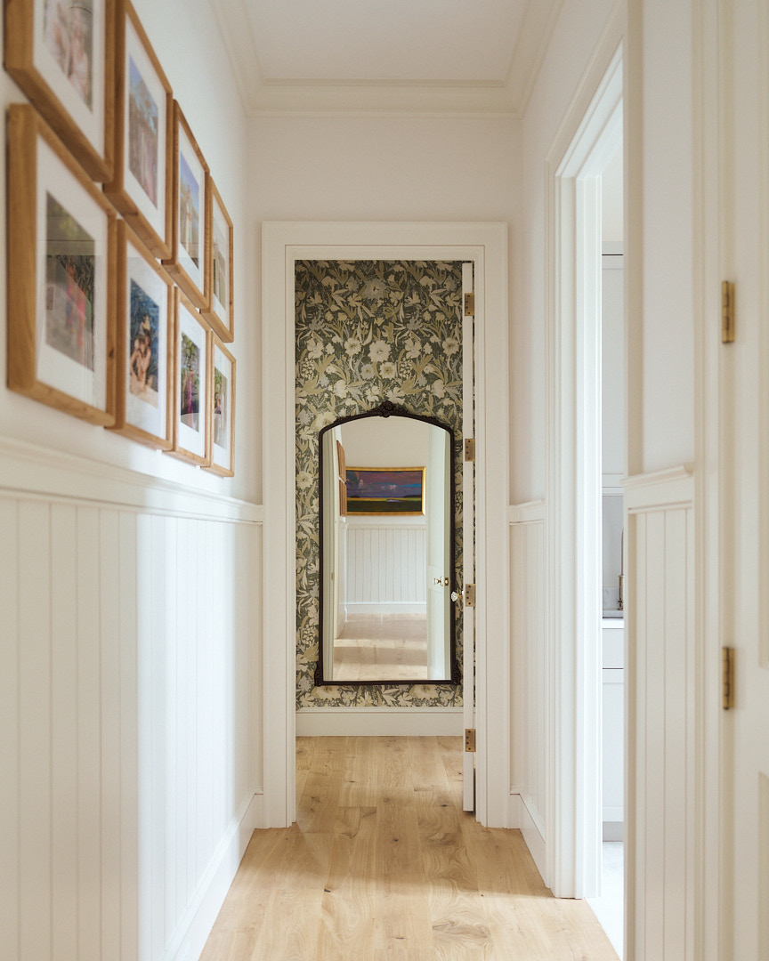 Bright hallway with custom millwork, gallery-style family photography, wide plank oak floors, and a statement mirror framed by botanical wallpaper.