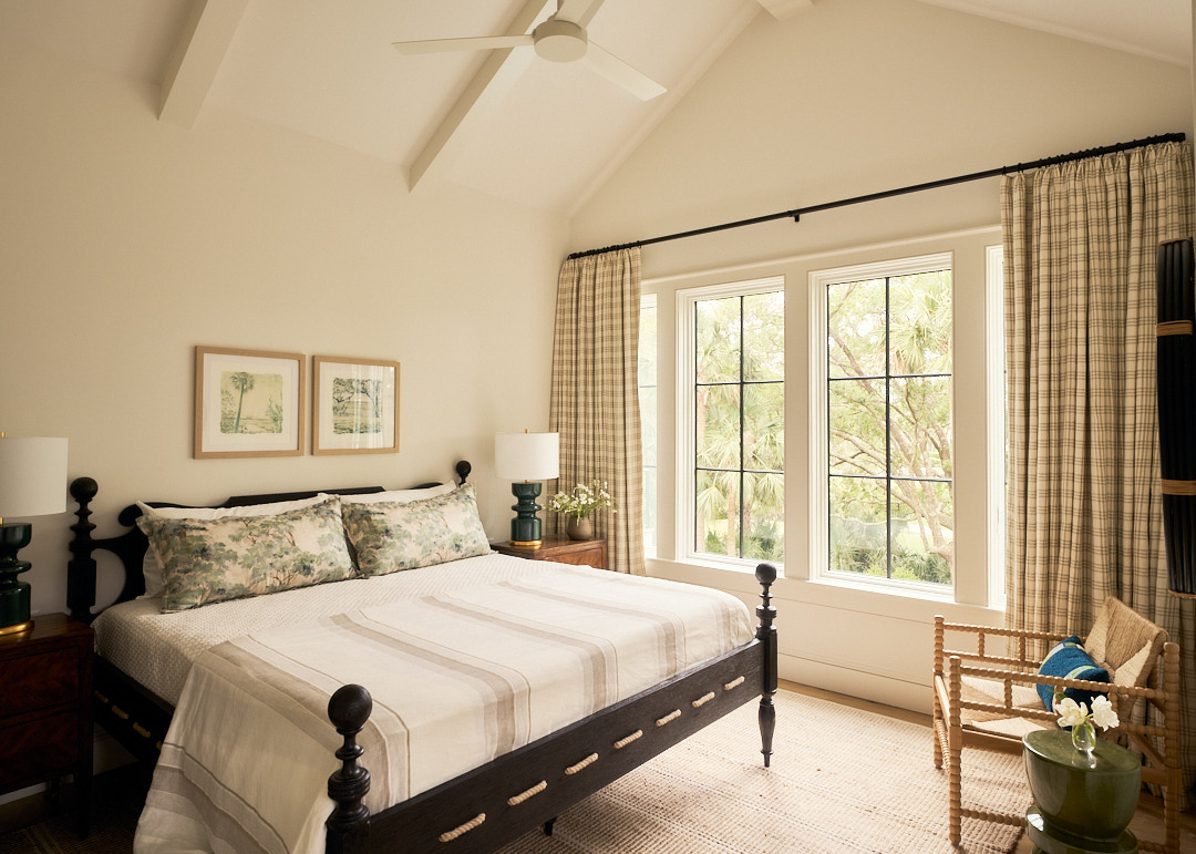 Inviting guest bedroom with a dark wood spindle bed, plaid drapery panels, woven accent chair, and expansive windows overlooking Kiawah Island’s natural landscape.