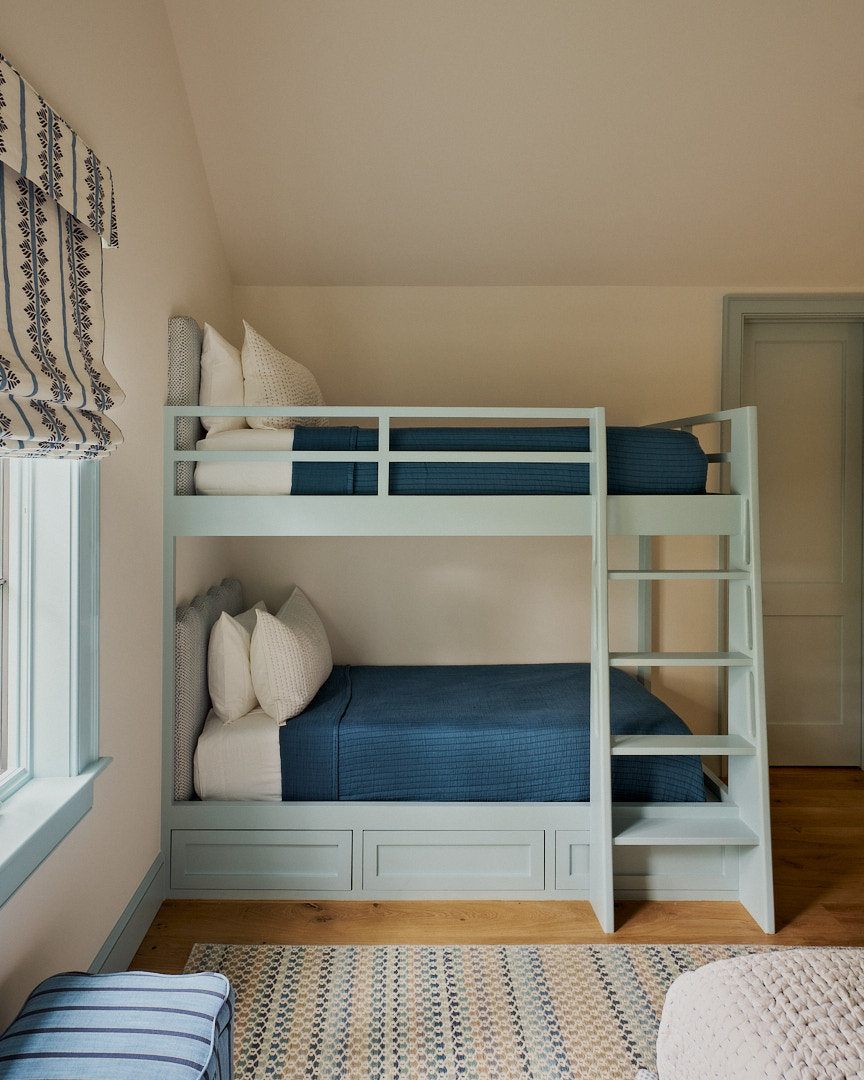 Custom bunk room featuring built-in pale blue bunk beds, integrated storage drawers, patterned Roman shades, and playful details designed for family gatherings.