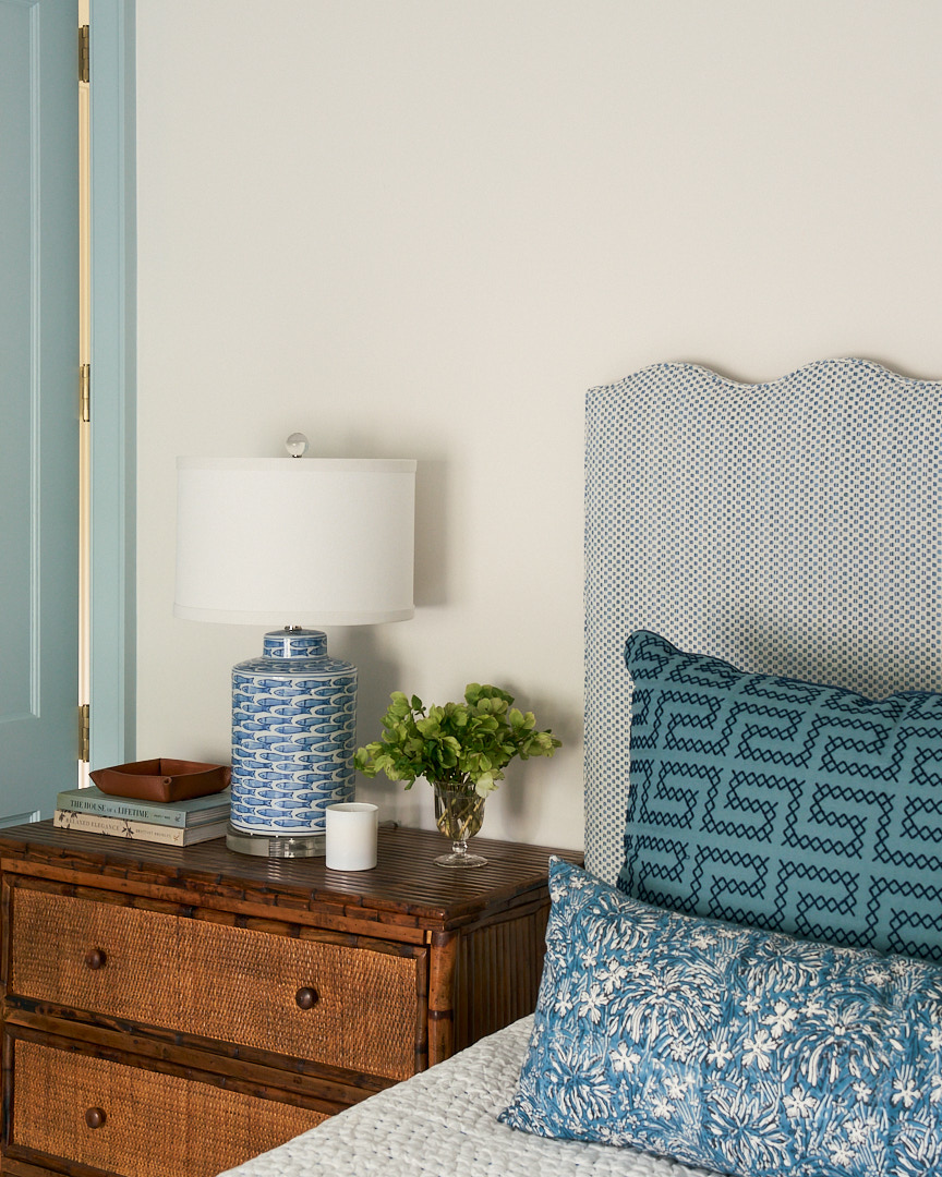 Guest bedroom vignette featuring a woven nightstand, blue-and-white ceramic lamp, upholstered headboard, and layered coastal textiles in a luxury Kiawah Island home.