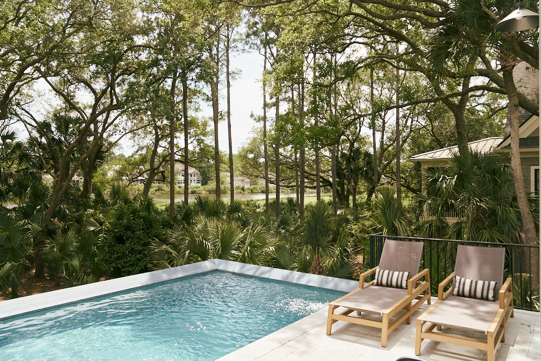 Luxury Kiawah Island backyard retreat featuring a modern swimming pool surrounded by mature live oaks, palmetto palms, and lush Lowcountry landscaping, with resort-style chaise lounges overlooking a scenic lagoon and golf course view.