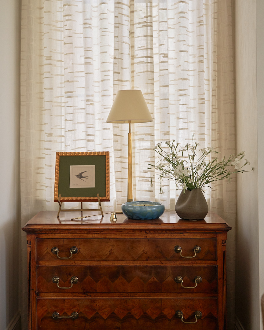 Antique walnut chest styled with framed botanical artwork, brass table lamp, and ceramic accessories beneath custom linen drapery in a luxury Kiawah Island residence by PWD Studio.