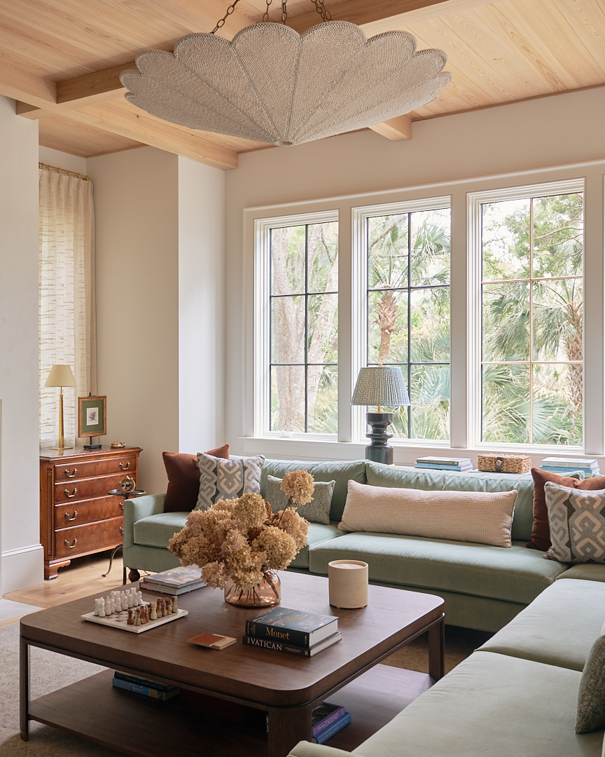 Light-filled Kiawah Island living room featuring a custom sage green sectional, sculptural statement chandelier, natural wood ceiling beams, and expansive windows overlooking the Lowcountry landscape.