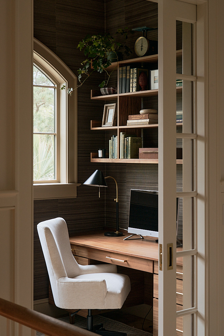 Custom home office with walnut built-in desk, floating wood shelves, dark grasscloth wallcovering, arched window, and tailored workspace designed for a luxury Kiawah Island residence.