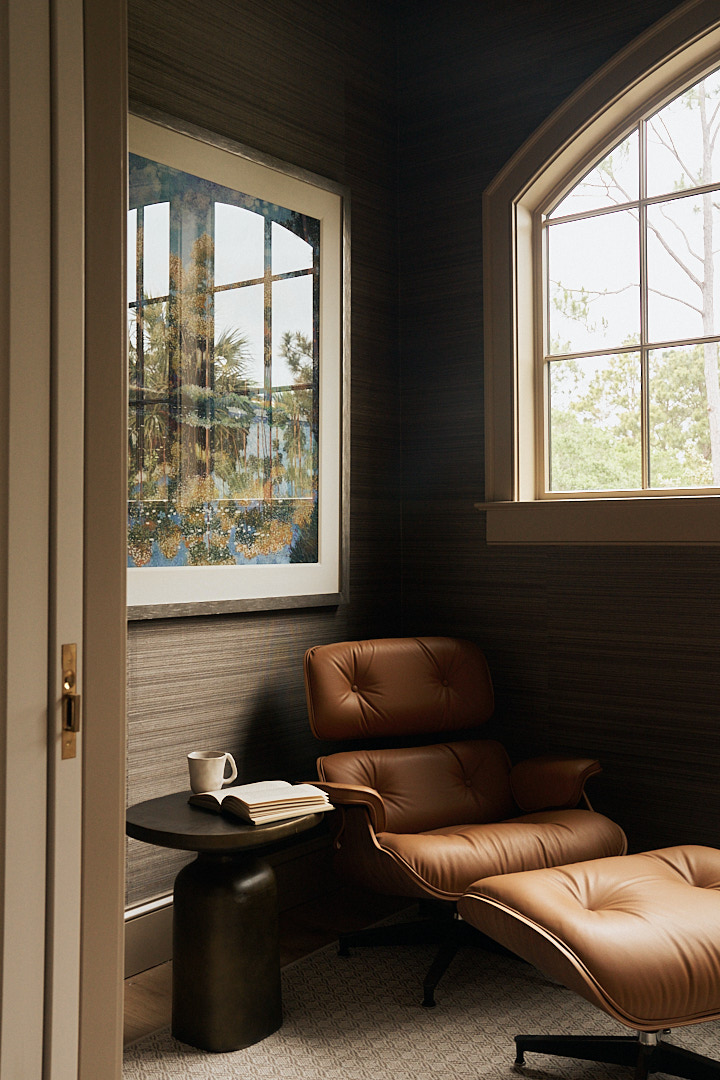 Moody study retreat featuring a classic leather lounge chair and ottoman, dark textured wallcovering, framed artwork, and an arched window overlooking the natural Lowcountry landscape.Bespoke home office with custom walnut cabinetry, floating bookshelves, 