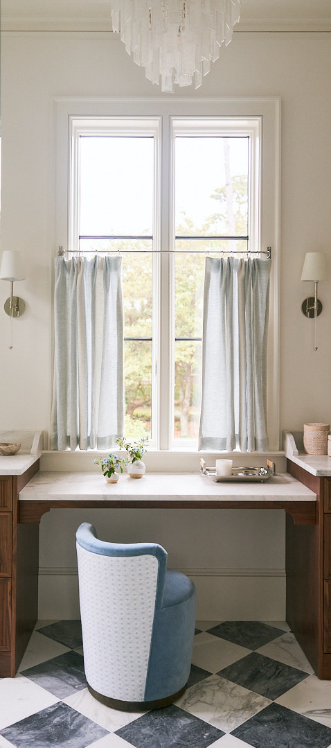 Built-in makeup vanity with marble countertop, upholstered blue chair, and large windows bringing natural light into a sophisticated Kiawah Island primary bath.