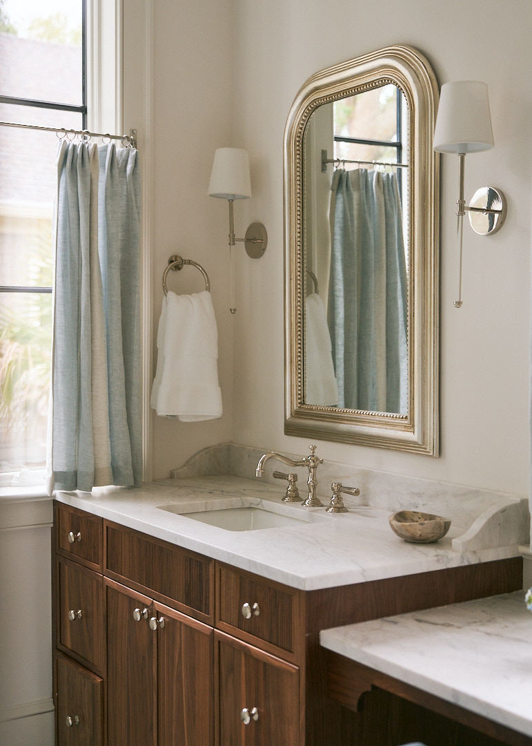 Custom walnut vanity with marble countertop, polished nickel fixtures, framed mirror, and tailored linen café curtains in an elegant coastal primary bathroom.