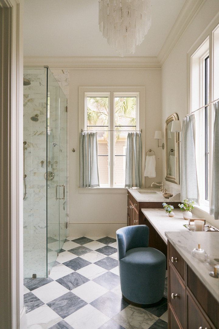 Luxury primary bathroom with walnut vanities, marble countertops, a glass-enclosed shower, checkerboard marble flooring, and soft blue accents overlooking tropical Lowcountry views.